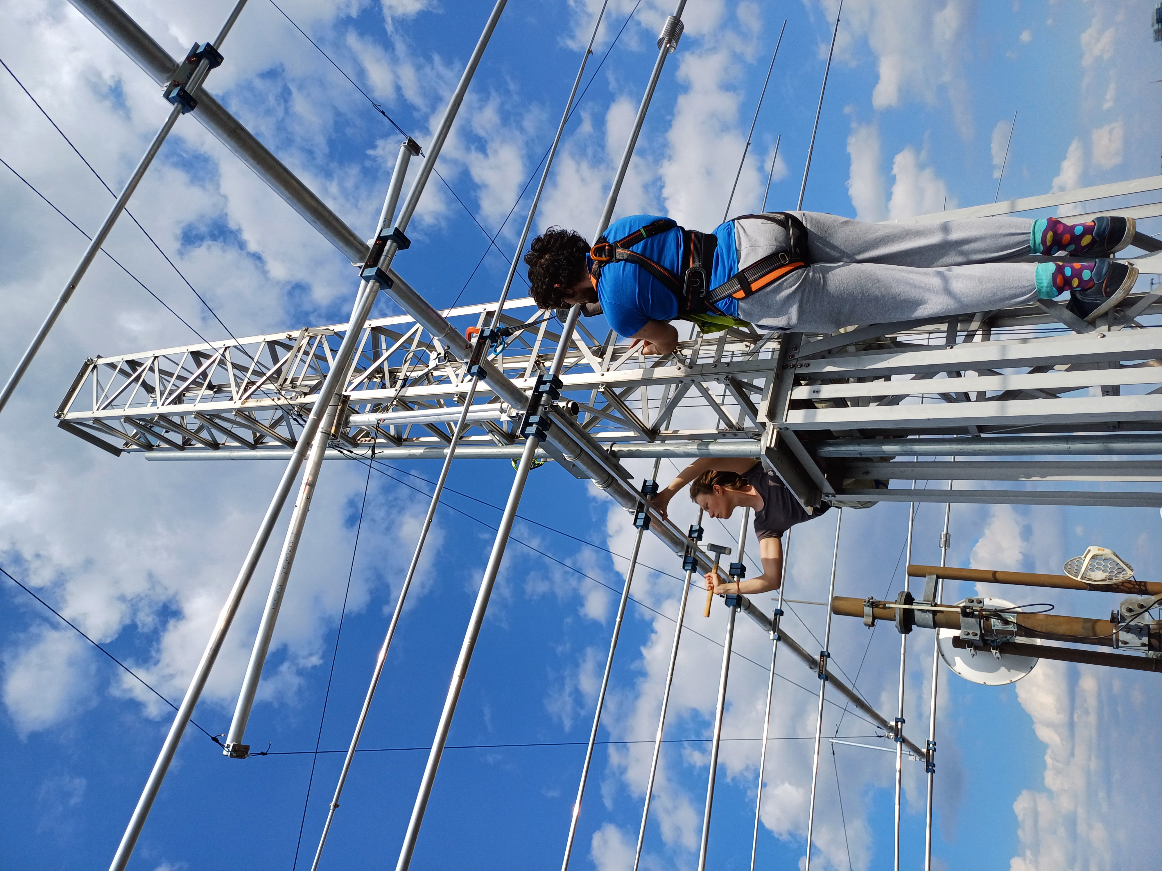 DL7OAR und DK3NN hängen in Kletterzeug unter dem Einspeisepunkt der Antenne. Im Hintergrund blauer Himmel mit Schäfchenwölkchen. DL7OAR arbeitet mit einem Gummihammer